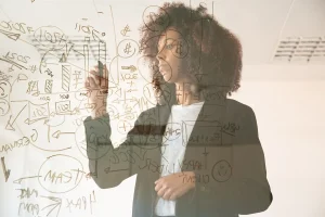Mulher com cabelo cacheado escrevendo em um vidro, com gráficos e anotações sobre planejamento e estratégia de negócios. Como impulsionar resultados e criatividade.