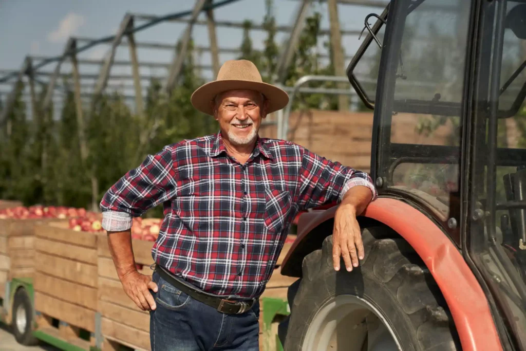 Homem idoso sorridente em fazenda, posando ao lado de um trator, com caixas de tomates ao fundo, vestindo chapéu de palha e roupa de fazendeiro, em ambiente rural.