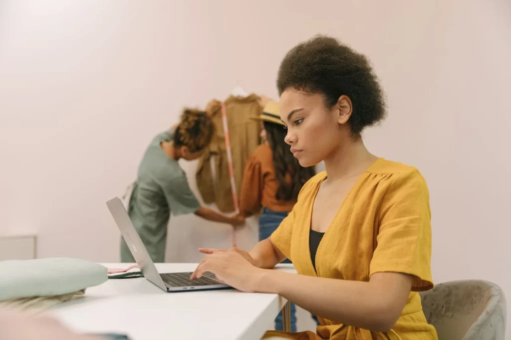 Mulher trabalhando em um laptop em um escritório, enquanto outras pessoas aparecem ao fundo. Cena de trabalho colaborativo que remete à Maturidade Digital das PMEs, com foco em avanço tímido ou ponto de partida.