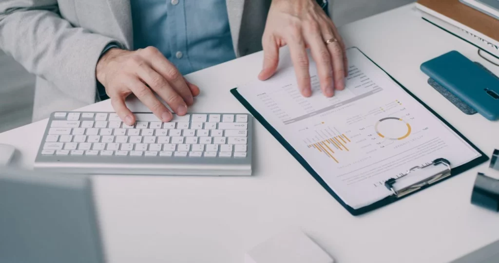 Pessoa em reunião usando teclado e analisando gráficos e relatórios em tablet no escritório, enquanto documentos e celular ficam sobre a mesa. Tecnologia e integração de dados para gestão empresarial.
