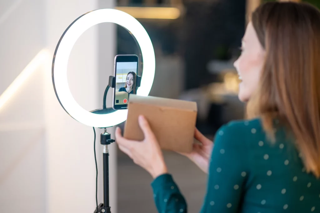 Mulher em estúdio usando microfone e anel de luz para gravar um vídeo enquanto conversa com um assistente de voz no celular; foco em Marketing de Voz: Otimizando Conteúdo para Assistentes e Buscas Por Voz
