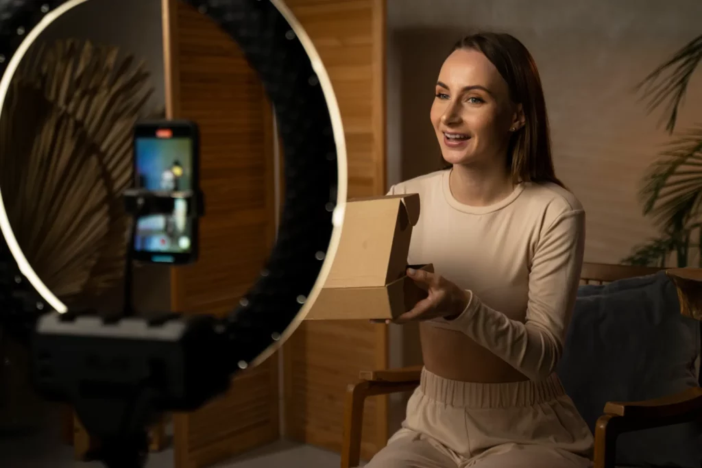 TikTok Shop no Brasil: O Que Aprender Com o Crescimento Acelerado da Plataforma: Pessoa em casa recebendo uma encomenda em uma caixa de papelão, em um ambiente iluminado, com celular sendo usado para gravar ou compartilhar o pedido em vídeo.