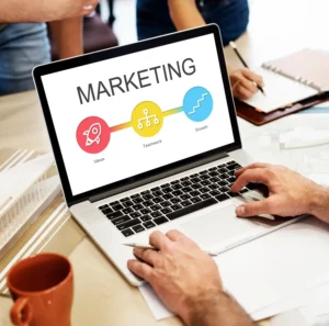 Como criar uma estratégia de marketing digital para pequenas empresas: notebook exibindo o termo MARKETING com gráfico em cores, enquanto pessoas analisam dados e fazem anotações em uma mesa de trabalho.