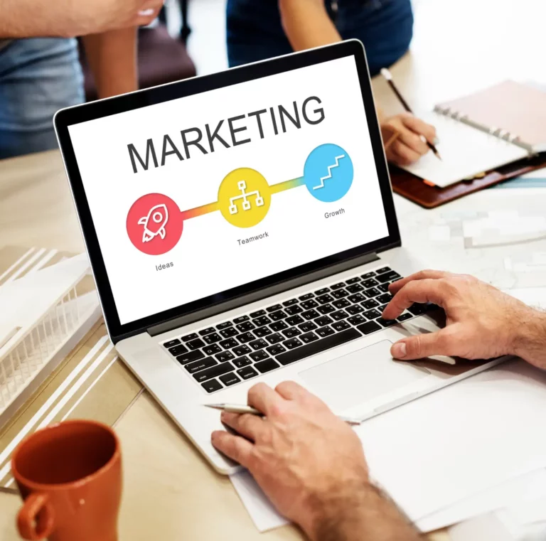 Como criar uma estratégia de marketing digital para pequenas empresas: notebook exibindo o termo MARKETING com gráfico em cores, enquanto pessoas analisam dados e fazem anotações em uma mesa de trabalho.