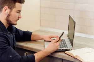 Presença Digital: O Que Sua Empresa Precisa Ter Para Ser Encontrada na Internet: Homem com fones de ouvido usando smartphone e trabalhando em um notebook em mesa de escritório, com livro aberto ao lado, em ambiente iluminado.