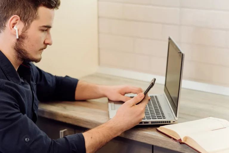 Presença Digital: O Que Sua Empresa Precisa Ter Para Ser Encontrada na Internet: Homem com fones de ouvido usando smartphone e trabalhando em um notebook em mesa de escritório, com livro aberto ao lado, em ambiente iluminado.
