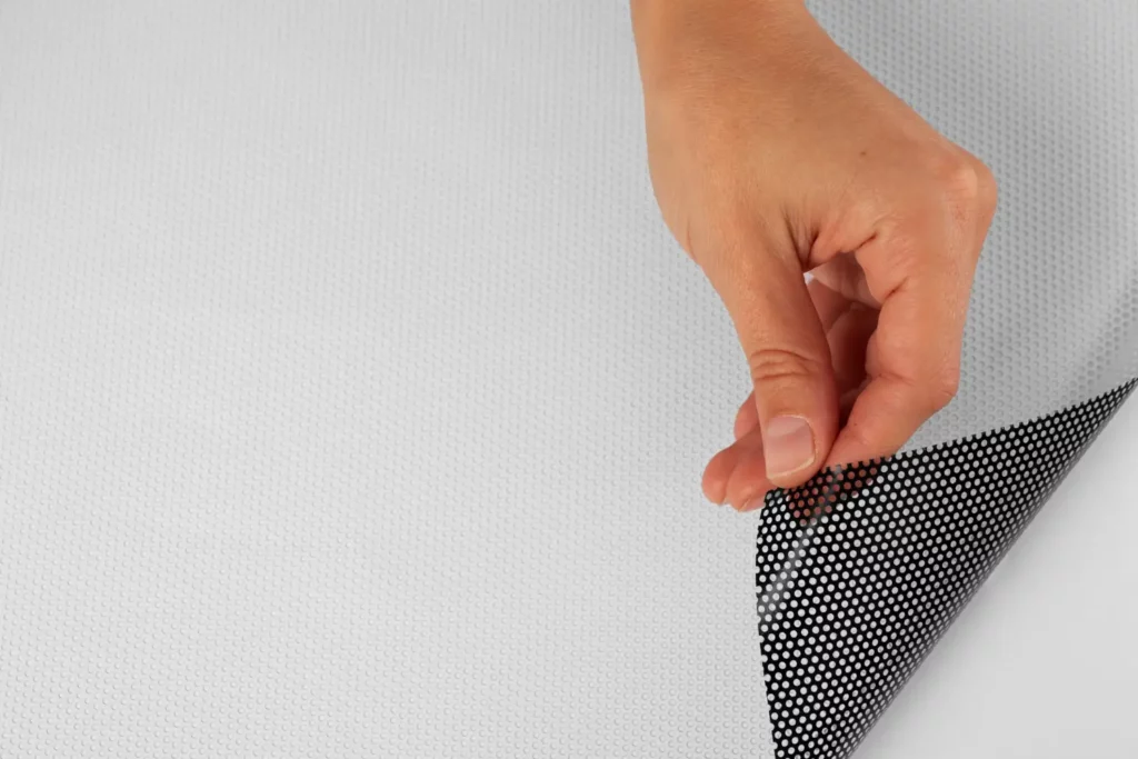 Manta de Polietileno: Onde Utilizar e Quais São as Vantagens Desse Material: Mão segurando e ajustando uma manta de polietileno com acabamento pontilhado antideslizante sobre uma superfície clara, evidenciando sua textura e aplicação como proteção